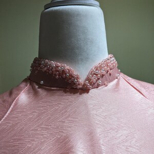 May include: Close-up of a pink dress with a high, beaded collar. The collar is adorned with small, light pink pearls. The dress has a subtle, textured pattern. The fabric appears to be a soft, satin-like material.