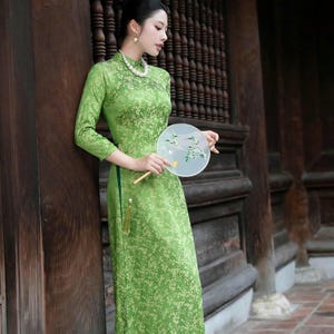 May include: A long, green floral print dress with long sleeves and a mandarin collar. The dress is accessorized with a pearl necklace and a decorative fan. The background features wooden architectural details.