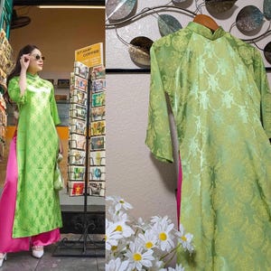 May include: A lime green, long-sleeved dress with a mandarin collar and a floral pattern. The dress is paired with bright pink pants. The dress is hanging on a wooden hanger.