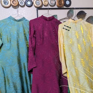 Op de afbeelding: Drie vintage jurken met mandarijnkragen hangen aan een muur. De jurken zijn teal, bordeauxrood en geel, elk met een bloemenpatroon. De gele jurk heeft zwarte kikker sluitingen. De jurken zijn gemaakt van een glanzende stof.
