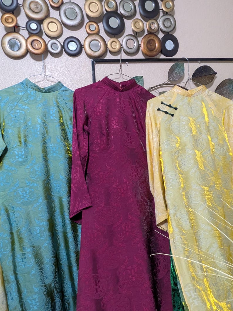 Op de afbeelding: Drie vintage jurken met mandarijnkragen hangen aan een muur. De jurken zijn teal, bordeauxrood en geel, elk met een bloemenpatroon. De gele jurk heeft zwarte kikker sluitingen. De jurken zijn gemaakt van een glanzende stof.