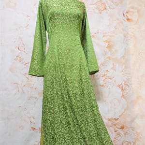 May include: A long, green dress with a floral pattern. The dress has long sleeves and a round neckline. The fabric has a subtle sheen. The dress is displayed on a mannequin against a floral background.