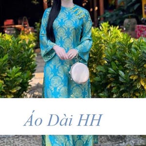 May include: A woman wearing a teal Áo Dài dress with a gold floral pattern. The dress has long sleeves and a high collar with gold accents. She is holding a small, round, silver purse. The text "Áo Dài HH" is at the bottom of the image.