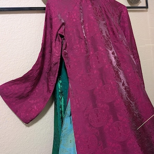Op de afbeelding: Een magenta jurk met lange mouwen en een mandarijnkraag, met een patroon. De jurk heeft een hoge halslijn en een vloeiend silhouet. Het is gelaagd over groene en blauwe stoffen.