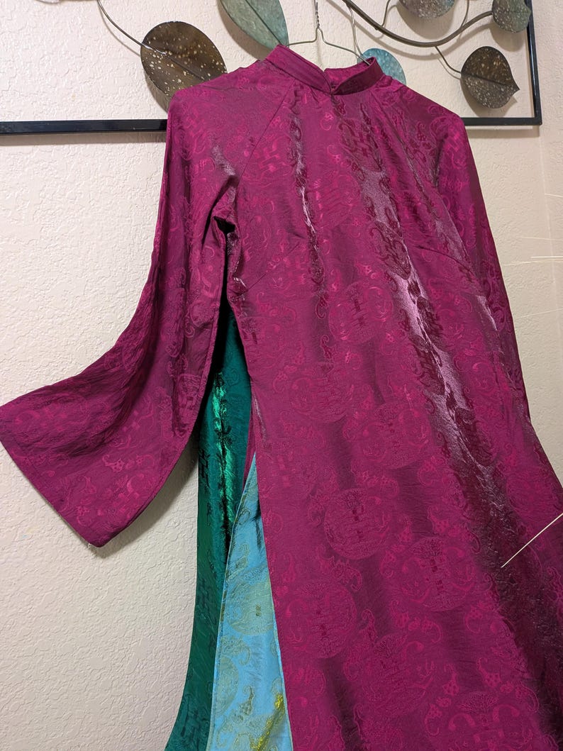 Op de afbeelding: Een magenta jurk met lange mouwen en een mandarijnkraag, met een patroon. De jurk heeft een hoge halslijn en een vloeiend silhouet. Het is gelaagd over groene en blauwe stoffen.