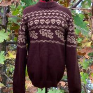 May include: A dark maroon long-sleeve sweater featuring a ribbed mock neck collar and a tan-colored horizontal band across the chest and shoulders. The band displays a repeating pattern of acorns and oak leaves with zigzag border detailing.