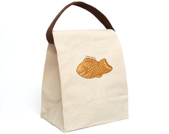 Puede incluir: Una bolsa de almuerzo de lona blanca con asas marrones. La bolsa tiene un gráfico de un taiyaki, un pastel japonés con forma de pez, en la parte delantera. El texto "Taiyaki" está impreso sobre el gráfico.