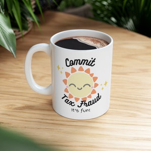 Puede incluir: Taza de cerámica blanca con asa negra. La taza tiene un sol de dibujos animados con una cara sonriente y el texto "Commit Tax Fraud It's fun!" impreso en ella.