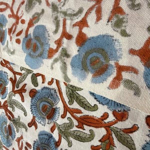 Op de afbeelding: Close-up van een stof met een herhalend bloemenpatroon. Het ontwerp toont blauwe bloemen met oranje en groene accenten op een cr&egrave;mekleurige achtergrond. De stof lijkt katoen of linnen te zijn, met een getextureerde weefsel.