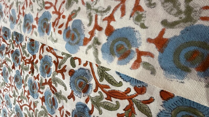 Op de afbeelding: Close-up van een stof met een herhalend bloemenpatroon. Het ontwerp toont blauwe bloemen met oranje en groene accenten op een cr&egrave;mekleurige achtergrond. De stof lijkt katoen of linnen te zijn, met een getextureerde weefsel.