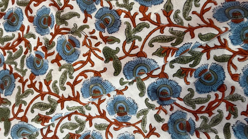 Op de afbeelding: Close-up van een stof met een herhalend bloemenpatroon. Het ontwerp heeft lichtblauwe bloemen met groene en bruine accenten op een witte achtergrond. Het bloemenpatroon is gedetailleerd en bedekt het hele oppervlak.