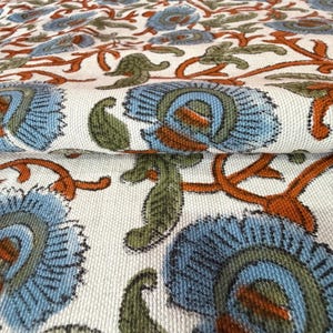 Op de afbeelding: Close-up van een gevouwen stof met een herhalend bloemenpatroon. Het ontwerp toont grote blauwe bloemen met oranje en groene accenten op een cr&egrave;mekleurige achtergrond. De stof heeft een getextureerde uitstraling, wat wijst op een natuurlijke vezel.