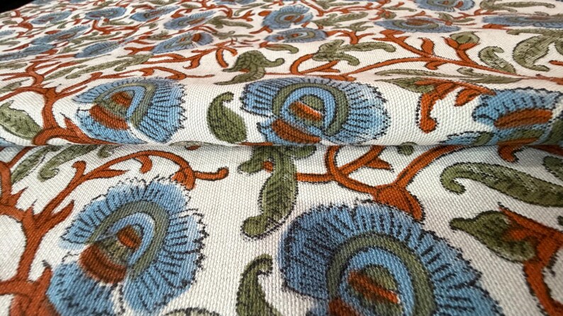 Op de afbeelding: Close-up van een gevouwen stof met een herhalend bloemenpatroon. Het ontwerp toont grote blauwe bloemen met oranje en groene accenten op een cr&egrave;mekleurige achtergrond. De stof heeft een getextureerde uitstraling, wat wijst op een natuurlijke vezel.