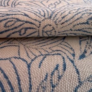 Op de afbeelding: Close-up van een gevouwen stof met een crèmekleurige basis en een herhalend patroon van blauwe bloemmotieven. De geweven stof toont het patroon op zowel de bovenste als de gevouwen lagen, met een getextureerde uitstraling.