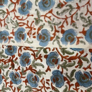 Op de afbeelding: Close-up van een stof met een bloemenpatroon. Het ontwerp toont lichtblauwe bloemen met bruine centra en stelen, en groene bladeren, tegen een cr&egrave;mekleurige achtergrond. De stof is waarschijnlijk katoen of linnen.