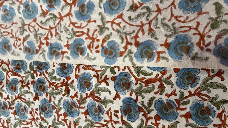 Op de afbeelding: Close-up van een stof met een bloemenpatroon. Het ontwerp toont lichtblauwe bloemen met bruine centra en stelen, en groene bladeren, tegen een cr&egrave;mekleurige achtergrond. De stof is waarschijnlijk katoen of linnen.