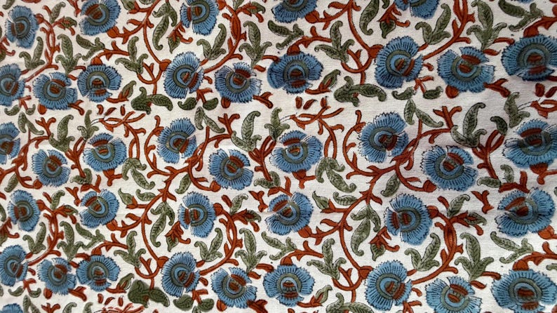 Op de afbeelding: Stof met een herhalend bloemenpatroon. Het ontwerp omvat blauwe bloemen, bruine stelen en groene bladeren tegen een witte achtergrond. Het patroon is gedetailleerd en symmetrisch.