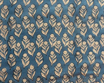 May include: A close-up of a blue fabric with a repeating pattern of stylized white floral designs. Each design features a central flower and surrounding leaf shapes, creating a textured appearance. The fabric has a slightly mottled look.