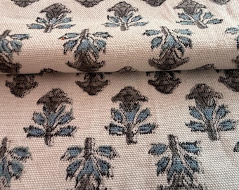 May include: Close-up of a folded fabric with a repeating pattern of stylized floral motifs. The design features blue and gray leaves on a cream-colored background. The fabric has a textured appearance, suggesting a woven material.