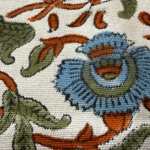 Op de afbeelding: Close-up van een stof met een bloemenpatroon. Het ontwerp toont blauwe bloemen met groene en oranje accenten op een cr&egrave;mekleurige achtergrond. De stof heeft een getextureerde uitstraling.