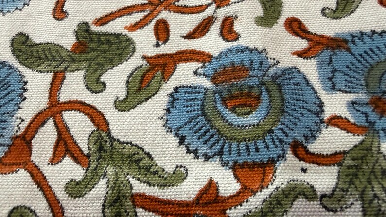 Op de afbeelding: Close-up van een stof met een bloemenpatroon. Het ontwerp toont blauwe bloemen met groene en oranje accenten op een cr&egrave;mekleurige achtergrond. De stof heeft een getextureerde uitstraling.