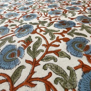 Op de afbeelding: Close-up van een stof met een bloemenpatroon. Het ontwerp heeft lichtblauwe bloemen met oranje en groene accenten op een cr&egrave;mekleurige achtergrond. De stof heeft een getextureerde uitstraling.