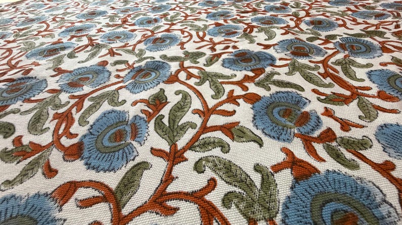 Op de afbeelding: Close-up van een stof met een bloemenpatroon. Het ontwerp heeft lichtblauwe bloemen met oranje en groene accenten op een cr&egrave;mekleurige achtergrond. De stof heeft een getextureerde uitstraling.
