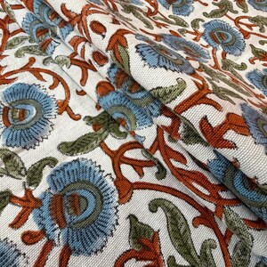 Op de afbeelding: Close-up van een gevouwen stof met een bloemenpatroon. Het ontwerp toont lichtblauwe bloemen, groene bladeren en roestkleurige stelen tegen een witte achtergrond. De stof lijkt katoen of linnen te zijn, geschikt voor woondecoratie of kleding.