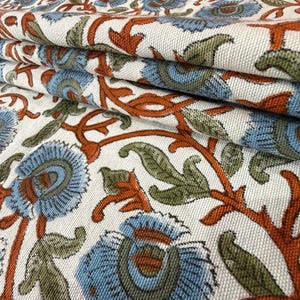 Op de afbeelding: Close-up van gevouwen stof met een bloemenpatroon. Het ontwerp toont blauwe bloemen met bruine centra en stelen, samen met groene bladeren, tegen een witte achtergrond. De stof is gelaagd en toont de gedetailleerde print.