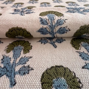 Puede incluir: Tela color crema con un patrón floral repetido en verde oliva y azul claro. La tela está doblada, mostrando la textura y el diseño de la impresión. Las flores tienen una apariencia detallada, dibujada a mano.