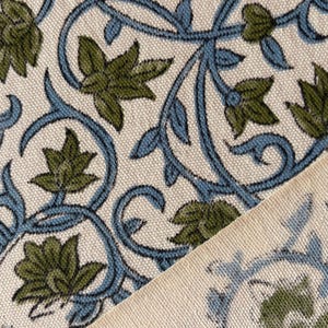 May include: Close-up of a fabric with a repeating floral pattern. The design features green flowers and blue vines on a cream-coloured background. The fabric is folded diagonally, revealing the pattern on both sides.
