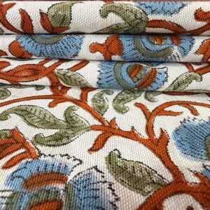 Op de afbeelding: Close-up van gevouwen stof met een herhalend bloemenpatroon. Het ontwerp heeft blauwe, oranje en groene bloemenelementen op een cr&egrave;mekleurige achtergrond. De stof is opgestapeld en toont de gedetailleerde print.