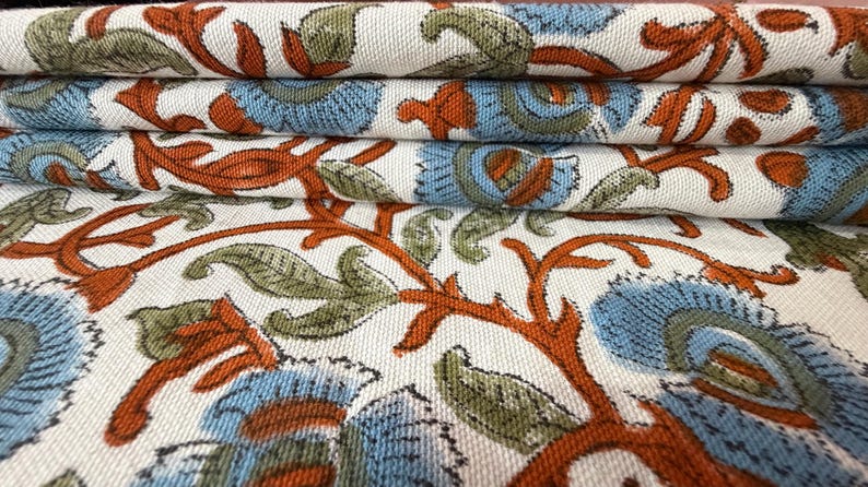Op de afbeelding: Close-up van gevouwen stof met een herhalend bloemenpatroon. Het ontwerp heeft blauwe, oranje en groene bloemenelementen op een cr&egrave;mekleurige achtergrond. De stof is opgestapeld en toont de gedetailleerde print.