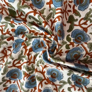 Op de afbeelding: Close-up van een witte stof met een herhalend bloemenpatroon. Het ontwerp heeft blauwe bloemen met bruine stelen en groene bladeren. De stof is gevouwen, waardoor schaduwen en textuur ontstaan.