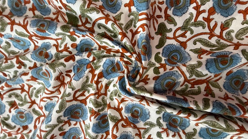 Op de afbeelding: Close-up van een witte stof met een herhalend bloemenpatroon. Het ontwerp heeft blauwe bloemen met bruine stelen en groene bladeren. De stof is gevouwen, waardoor schaduwen en textuur ontstaan.