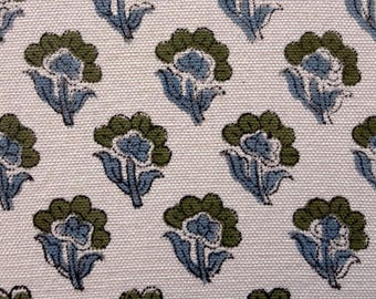 May include: A white fabric featuring a repeating pattern of small floral motifs. Each flower consists of a light blue center with dark blue outlines and a rounded olive green outer petal layer, arranged in a consistent grid across the textile.