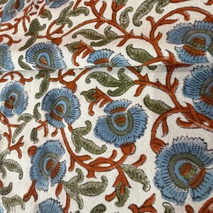 Op de afbeelding: Close-up van een witte stof met een herhalend bloemenpatroon. Het ontwerp toont grote blauwe bloemen met oranje centra en stelen, samen met groene bladeren. De stof is gevouwen en toont de continu&iuml;teit van het patroon.