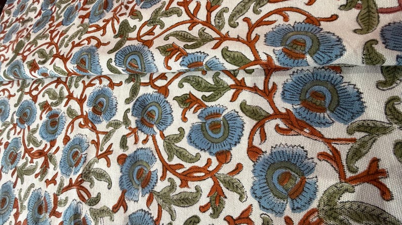 Op de afbeelding: Close-up van een witte stof met een herhalend bloemenpatroon. Het ontwerp toont grote blauwe bloemen met oranje centra en stelen, samen met groene bladeren. De stof is gevouwen en toont de continu&iuml;teit van het patroon.