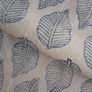 May include: Close-up of a beige fabric with a repeating pattern of blue leaf designs. The leaves have detailed veining and are printed on a textured, woven material. The fabric is folded, showing the pattern's continuity.