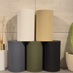 Könnte beinhalten: Fünf zylindrische Behälter mit gerippter Textur, pyramidenförmig gestapelt. Die Behälter sind weiß, beige, dunkelgrau, olivgrün und schwarz. Eine weiße Diffuserflasche mit goldenem Deckel und eine Topfpflanze mit Schlangenpflanze befinden sich im Hintergrund.