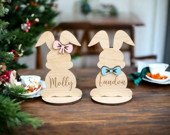 Op de afbeelding: Twee houten konijnvormige naambordhouders. Eén heeft een roze strik en de naam "Molly" gegraveerd. De andere heeft een blauwe strik en de naam "Landon". De konijnen staan op een donkere houten tafel.