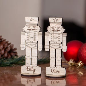 May include: Two wooden nutcracker figurines with star-topped hats and mustaches. The figurines are engraved with the names "Billy" and "Olive" on their bases. Red Christmas ornaments and a pinecone are in the background.
