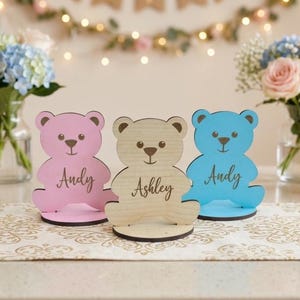 Könnte beinhalten: Drei bärenförmige Holzornamente auf runden Sockeln. Der linke Bär ist hellrosa mit dem Namen Andy, der mittlere Bär ist aus naturfarbenem Holz mit dem Namen Ashley und der rechte Bär ist hellblau mit dem eingravierten Namen Andy in Schreibschrift.