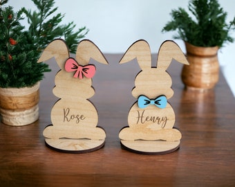 Op de afbeelding: Twee houten konijnenfiguren met gepersonaliseerde namen, Rose en Henry. Het konijn links heeft een roze strik en het konijn rechts een blauwe strik.