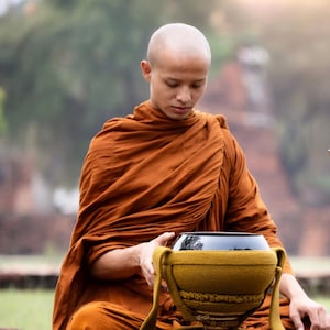 May include: A person in orange robes sits cross-legged, holding a black bowl with a yellow woven container. The person has a shaved head. The scene is set outdoors, with a blurred background of greenery and brick.