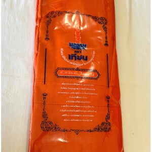 May include: A bright orange fabric cloth packaged in a clear plastic bag. The bag has a label with a black border and ornate design. The label features a blue and white logo with the text "ตรา เทียน" and "เครื่องหมาน" in Thai. The label also includes the text "เข้าทร ชีวร สบง ไเกมสวัส" in Thai.