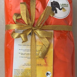May include: An orange package wrapped in clear cellophane and tied with a gold ribbon and bow. A circular sticker with an elephant logo and text is visible. The package contains a card with text.