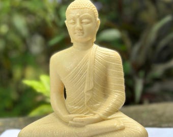 May include: A small, cream-colored statue of Buddha sitting in meditation pose. The statue is made of a smooth, slightly textured material and has a serene expression on its face.