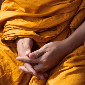 May include: A person's hands are folded together in their lap, covered by a bright orange robe. The robe is draped over the person's legs, creating a soft, flowing look.