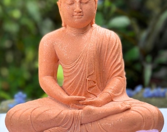 May include: A terracotta colored Buddha statue sitting in a meditative pose with closed eyes. The statue is sitting on a white surface with pink flowers in the foreground.